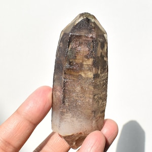May include: A natural smoky quartz crystal point. The crystal is a translucent brown color with a faceted top and a rough, textured surface. The crystal is held in a hand against a white background.