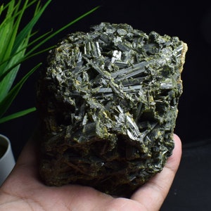 Könnte beinhalten: Ein großer, dunkelgrüner, grob strukturierter Kristallcluster. Die Kristalle sind lang und dünn, mit einem metallischen Glanz.