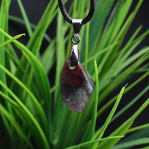 May include: A silver pendant necklace with a multi-coloured gemstone. The gemstone is a deep purple, red, and black colour. The necklace is hanging from a black cord.