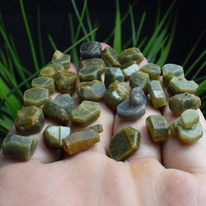 May include: A collection of green and grey gemstone rough, with a variety of shapes and sizes. The rough is displayed on a hand.