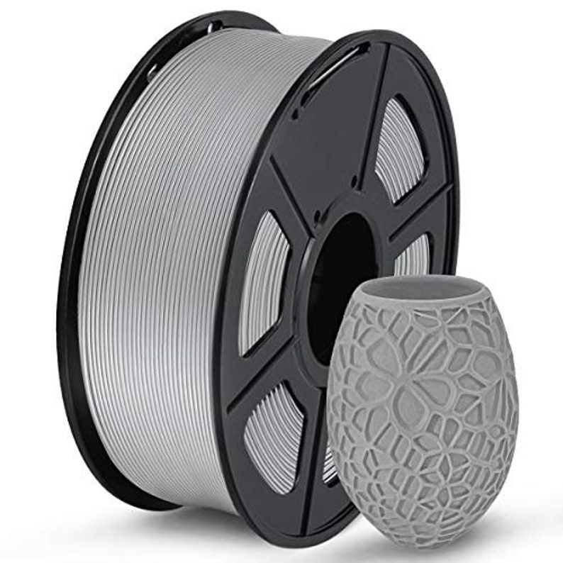 Pu&ograve; includere: Un rotolo di filamento color argento per la stampa 3D. Il filamento &egrave; avvolto attorno a un rotolo di plastica nera con un foro centrale. Il rotolo &egrave; su una superficie bianca.