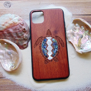 May include: A wooden phone case with a carved turtle design. The turtle's shell is made of iridescent abalone shell. The case is a dark reddish brown color.