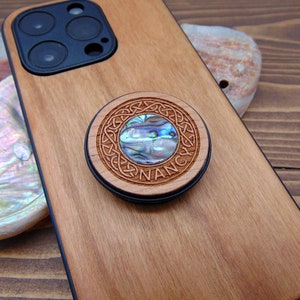 Op de afbeelding: Een houten telefoonhoesje met een zwarte cameralens en een ronde, houten telefoonhouder met een parelmoer inleg en het woord "NANCY" gegraveerd rond de rand.