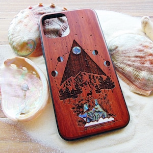 May include: A wooden phone case with a carved design of a mountain range, trees, and a crescent moon with stars. The case is made of dark red wood and has a natural wood finish. The design is inlaid with mother-of-pearl accents.