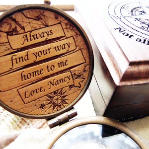 May include: A wooden compass with the engraved message "Always find your way home to me Love, Nancy." The compass is open, revealing a map design. A wooden box with a compass rose design and the text "Not all who wander are lost" is in the background.