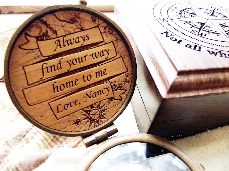 May include: A wooden compass with the engraved message "Always find your way home to me Love, Nancy." The compass is open, revealing a map design. A wooden box with a compass rose design and the text "Not all who wander are lost" is in the background.