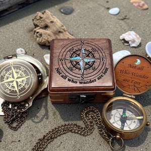 May include: A collection of vintage-style compasses and a wooden box with compass rose designs. One compass is open, displaying the directional needle. The box and a compass lid feature the quote, "Not all who wander are lost."