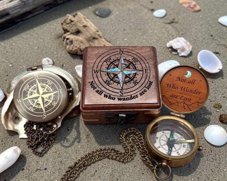 May include: A collection of vintage-style compasses and a wooden box with compass rose designs. One compass is open, displaying the directional needle. The box and a compass lid feature the quote, "Not all who wander are lost."