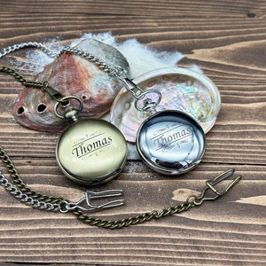 May include: Two round pocket watches with attached metal chains. One watch has a matte brass finish and the other has a polished silver finish. Both watches are engraved with the name Thomas in a script font with decorative flourishes above and below.