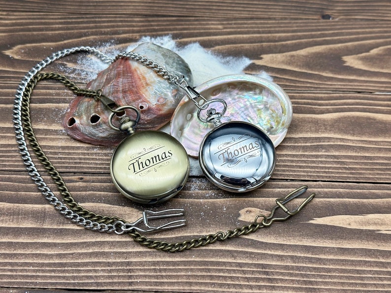 May include: Two round pocket watches with attached metal chains. One watch has a matte brass finish and the other has a polished silver finish. Both watches are engraved with the name Thomas in a script font with decorative flourishes above and below.
