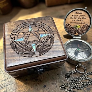 May include: A square wooden box with a compass design on the lid featuring the text TO THINE OWN SELF BE TRUE, UNITY, SERVICE, RECOVERY and iridescent inlays. Beside it, an open silver pocket compass with a chain and engraved lid text.