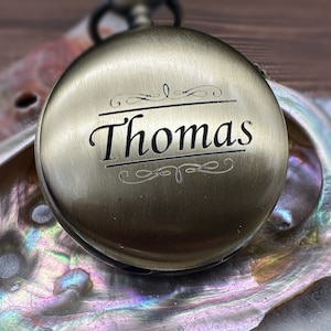 May include: A round, antique brass-toned pocket watch with a smooth, metallic finish. The front cover is engraved with the name Thomas in a serif font, centered between two decorative scrollwork lines. The watch features a small top winding crown.