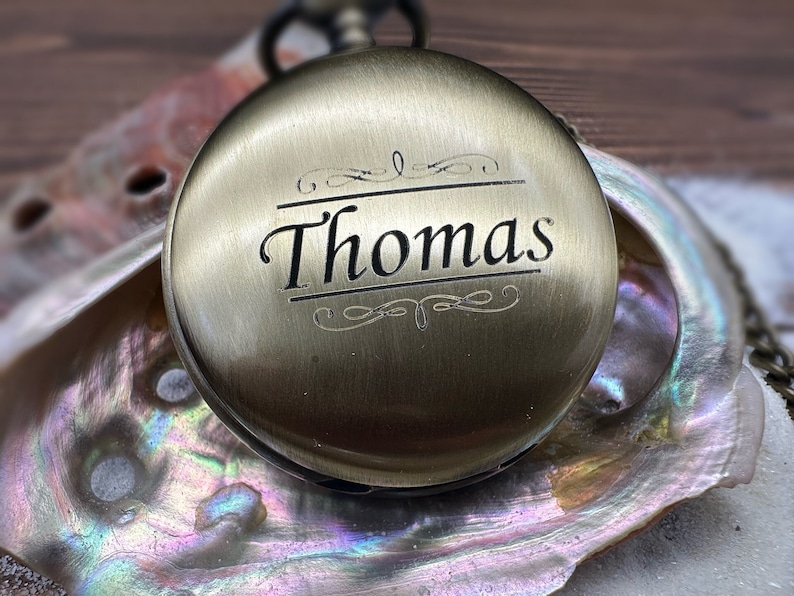 May include: A round, antique brass-toned pocket watch with a smooth, metallic finish. The front cover is engraved with the name Thomas in a serif font, centered between two decorative scrollwork lines. The watch features a small top winding crown.