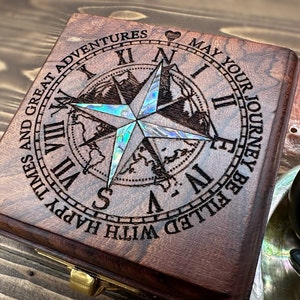 May include: A square dark brown wooden box featuring an engraved compass rose with iridescent abalone shell inlay. The lid is inscribed with the text: MAY YOUR JOURNEY BE FILLED WITH HAPPY TIMES AND GREAT ADVENTURES, surrounding Roman numerals.