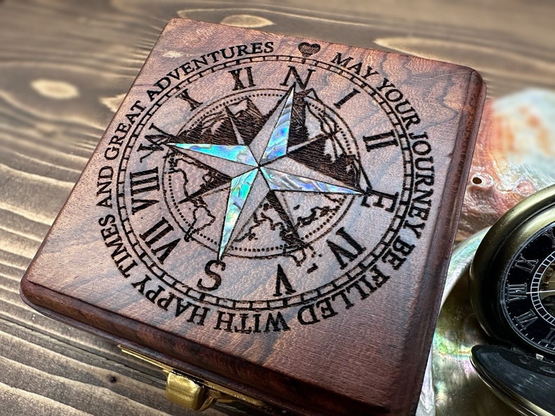 May include: A square dark brown wooden box featuring an engraved compass rose with iridescent abalone shell inlay. The lid is inscribed with the text: MAY YOUR JOURNEY BE FILLED WITH HAPPY TIMES AND GREAT ADVENTURES, surrounding Roman numerals.