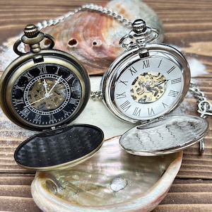 May include: Two mechanical pocket watches with chains. One watch has a bronze-toned case with a black dial and Roman numerals. The other watch has a silver-toned case with a white dial and Roman numerals. Both feature open centers revealing internal gears.