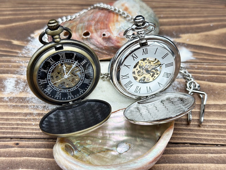 May include: Two mechanical pocket watches with chains. One watch has a bronze-toned case with a black dial and Roman numerals. The other watch has a silver-toned case with a white dial and Roman numerals. Both feature open centers revealing internal gears.