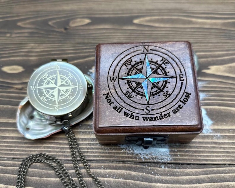 May include: A vintage-style compass and wooden box with a compass rose design. The box is engraved with the phrase "Not all who wander are lost." The compass is silver-toned with a chain. The box has a mother-of-pearl inlay.