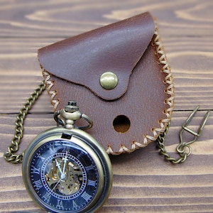 May include: A bronze-toned mechanical pocket watch with a black face, Roman numerals, and an exposed gear movement, attached to a matching metal chain. It is paired with a small, brown leather pouch featuring a brass snap closure and stitching.