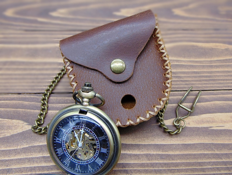 May include: A bronze-toned mechanical pocket watch with a black face, Roman numerals, and an exposed gear movement, attached to a matching metal chain. It is paired with a small, brown leather pouch featuring a brass snap closure and stitching.