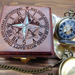 May include: A square wooden box with a laser-engraved compass rose and Roman numerals, featuring an iridescent inlay. The text reads: MAY YOUR JOURNEY BE FILLED WITH HAPPY TIMES AND GREAT ADVENTURES. Two brass-toned pocket watches are nearby.