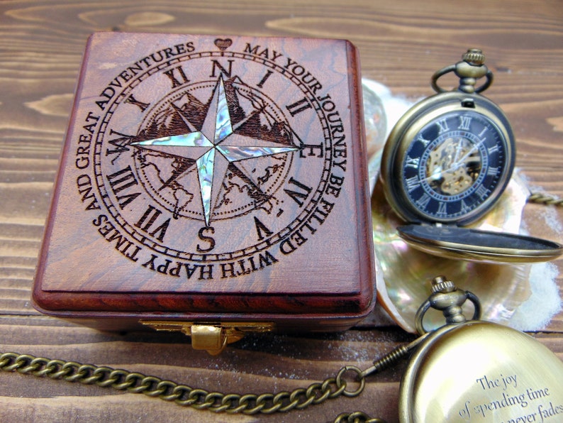 May include: A square wooden box with a laser-engraved compass rose and Roman numerals, featuring an iridescent inlay. The text reads: MAY YOUR JOURNEY BE FILLED WITH HAPPY TIMES AND GREAT ADVENTURES. Two brass-toned pocket watches are nearby.