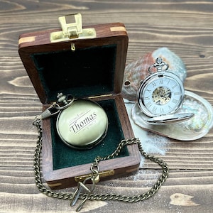 May include: A brass-toned pocket watch engraved with the name Thomas, resting inside a small wooden box with a dark green velvet lining. A silver-toned pocket watch with a white face and Roman numerals sits open on a nearby iridescent seashell.
