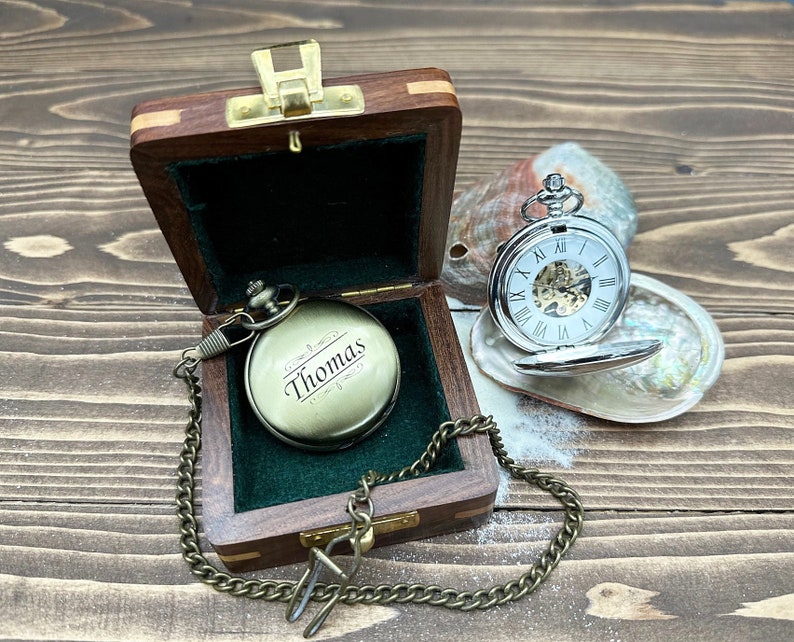 May include: A brass-toned pocket watch engraved with the name Thomas, resting inside a small wooden box with a dark green velvet lining. A silver-toned pocket watch with a white face and Roman numerals sits open on a nearby iridescent seashell.