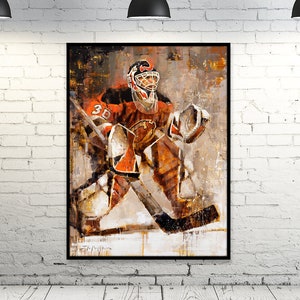 May include: An abstract painting of a hockey goalie in orange and white gear, set against a neutral background. The artwork is framed in black and hangs on a white brick wall, illuminated by three black pendant lights.