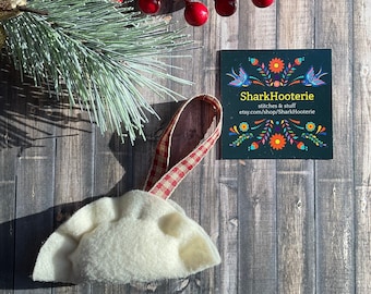 May include: White felt ornament shaped like a dumpling with a red and white gingham ribbon. The ornament is hanging from a wooden surface with a red berry sprig and a business card that reads "SharkHooterie stitches & stuff etsy.com/shop/SharkHooterie".