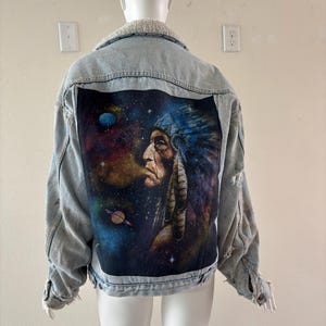 May include: Light blue denim jacket with a cream-colored shearling collar. The back features a vibrant graphic of a Native American chief against a cosmic background with planets and stars. The jacket has distressed details.