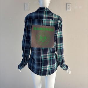 May include: A long-sleeved plaid shirt in shades of blue, teal, and white, displayed on a mannequin. The back of the shirt features a rectangular patch with the word "RAMONES" and a green emblem.
