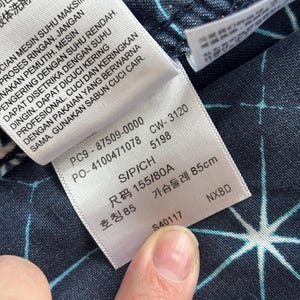 May include: Close-up of a clothing tag with text in multiple languages. The tag is white with black text and is attached to a dark blue fabric with a white starburst pattern. The tag includes size information.