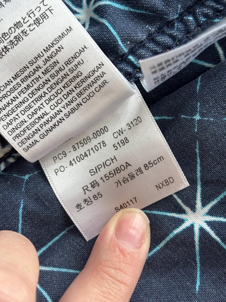 May include: Close-up of a clothing tag with text in multiple languages. The tag is white with black text and is attached to a dark blue fabric with a white starburst pattern. The tag includes size information.