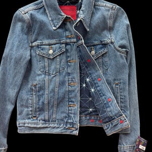 May include: A light blue denim jacket with a classic collar and button closure. The jacket features two chest pockets and a red label on the inside collar. The interior lining is navy blue with a white star pattern and red button accents.