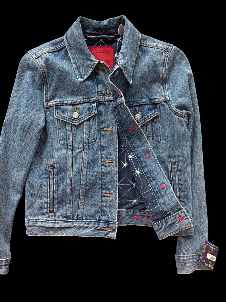 May include: A light blue denim jacket with a classic collar and button closure. The jacket features two chest pockets and a red label on the inside collar. The interior lining is navy blue with a white star pattern and red button accents.