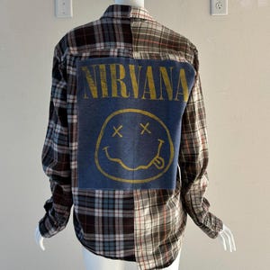 May include: A long-sleeved, button-down shirt with a patchwork design. The shirt features a blue denim panel on the back with the word "NIRVANA" in yellow lettering and a yellow smiley face graphic. The plaid pattern includes brown, gray, and red.