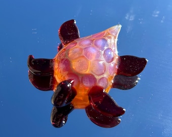 May include: A handcrafted glass turtle figurine with a vibrant color palette. The turtle's body is a mix of orange and pink hues, with a textured surface. Its flippers are a deep burgundy color, creating a striking contrast. The figurine is displayed against a reflective blue background.