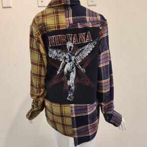 May include: A long-sleeve button-down shirt featuring a patchwork design of olive green and navy blue plaid flannel fabric. The back panel displays a large black rectangular patch with a white graphic of an angel and the text NIRVANA.