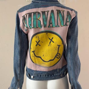 May include: A blue denim jacket with a pink back panel featuring the band name "NIRVANA" in teal lettering above a yellow smiley face with crossed-out eyes and a curved mouth. The jacket has a classic collar and button closures.