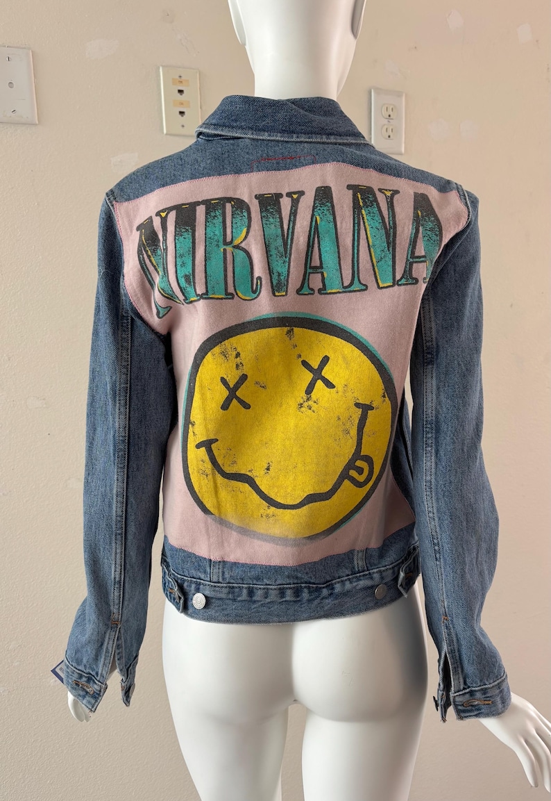 May include: A blue denim jacket with a pink back panel featuring the band name "NIRVANA" in teal lettering above a yellow smiley face with crossed-out eyes and a curved mouth. The jacket has a classic collar and button closures.