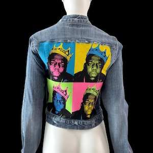 May include: A light blue denim jacket featuring a pop art style graphic of a person wearing a crown. The graphic is divided into four squares with vibrant colors: blue, yellow, green, and pink. The jacket has long sleeves and a classic collar.