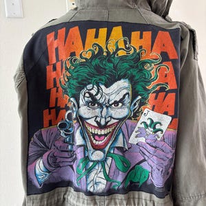 May include: An olive green jacket with a large graphic of the Joker on the back. The Joker is depicted with green hair, a purple suit, and a playing card. The word "HAHAHA" is printed in orange.