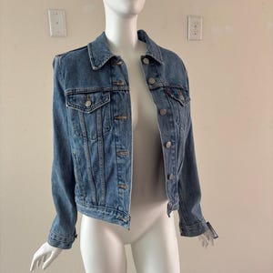 May include: A light blue denim jacket with silver-tone buttons and two chest pockets. The jacket is displayed on a mannequin. The jacket has a classic collar and long sleeves.