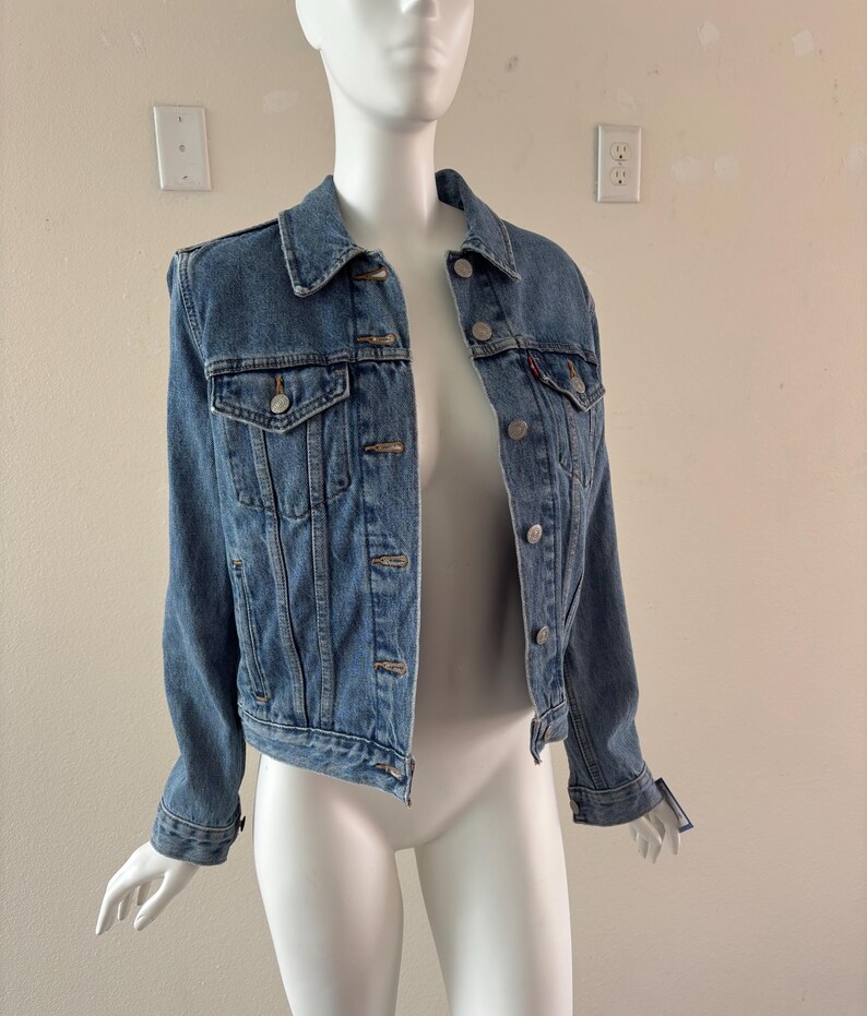 May include: A light blue denim jacket with silver-tone buttons and two chest pockets. The jacket is displayed on a mannequin. The jacket has a classic collar and long sleeves.