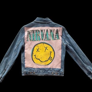 May include: Blue denim jacket with a pink back panel featuring the word "NIRVANA" in teal lettering above a yellow smiley face with crossed-out eyes and a protruding tongue. The jacket has a classic collar and button closures.