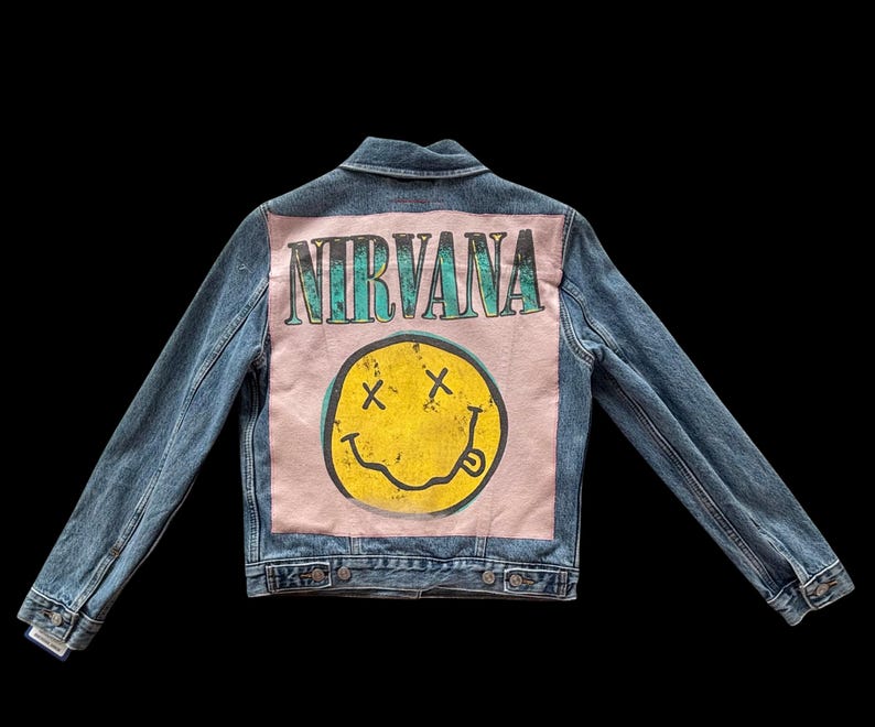 May include: Blue denim jacket with a pink back panel featuring the word "NIRVANA" in teal lettering above a yellow smiley face with crossed-out eyes and a protruding tongue. The jacket has a classic collar and button closures.