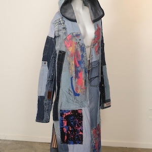 May include: A long, blue denim coat with a hood. The coat is made of patchwork denim and has a variety of colorful paint splatters and designs. The coat is open in the front and has a silver chain hanging from the neckline.