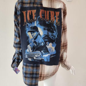 May include: A patchwork flannel shirt with a graphic of Ice Cube on the back. The shirt features a mix of blue, brown, and tan plaid patterns. The graphic is on a navy blue background and includes the text "ICE CUBE".