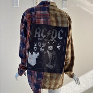May include: A long-sleeved flannel shirt with a patchwork design. The shirt features a blue, red, and brown plaid pattern on the sleeves and a brown and tan plaid pattern on the back. A black graphic of the band AC/DC with the text "HIGHWAY TO HELL" is on the back.